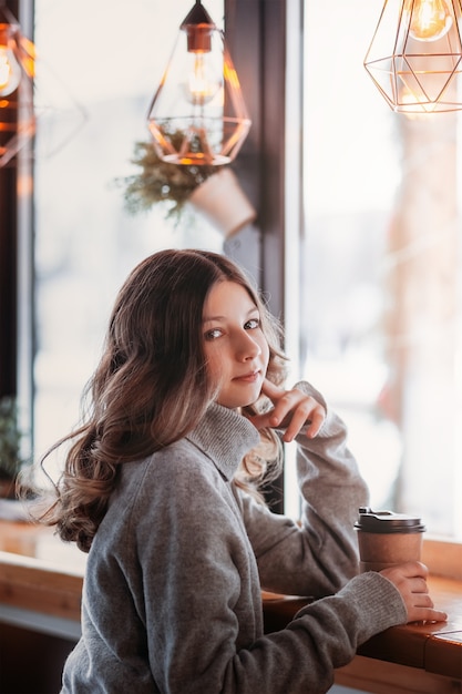 Молодая красивая девушка сидит в кафе с бумажным стаканчиком кофе