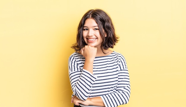 Young pretty girl smiling, enjoying life, feeling happy, friendly, satisfied and carefree with hand on chin