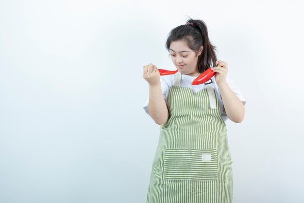 Young pretty girl standing in checkered apron and holding two red hot chili peppers .