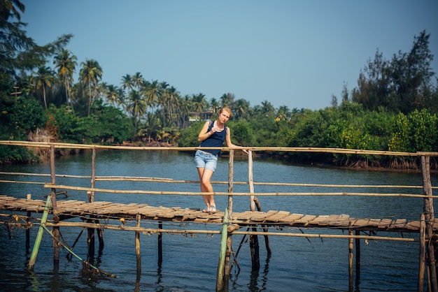 Young pretty girl stands on wooden bridge over small river against palm jungle and blue sky.