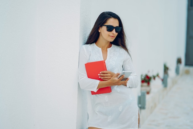 молодая и красивая девушка в летнем городе, стоя рядом с белой стеной с красной книгой и телефоном