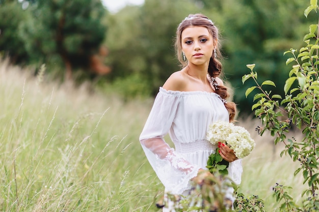 La giovane ragazza graziosa con il fiore in condimento leggero cammina a prato inglese vicino alla foresta dell'estate