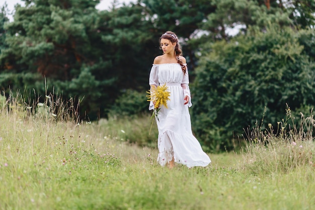 Young pretty girl with flower in light dressing walk at lawn near summer forest