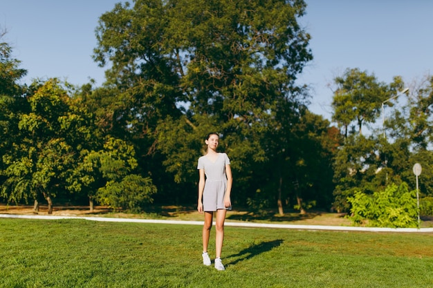 La giovane ragazza graziosa con capelli castani lunghi si è vestita in vestiti leggeri che rimangono sull'erba del prato inglese verde nel parco sullo sfondo degli alberi. tempo di sole estivo.