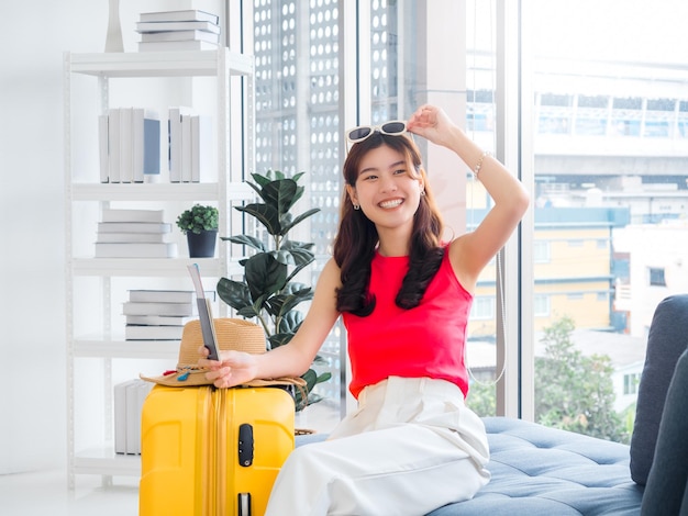 Young pretty happy Asian woman touching sunglasses on head holding passport with smiling sit on sofa near beach hat yellow suitcase in white living room ready to travel on summer vacation concept