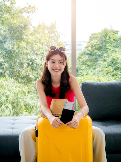 Young pretty happy asian woman with sunglasses holding passport and flight ticket with smiling sit on sofa near beach hat yellow suitcase vertical style ready to travel on summer vacation concept