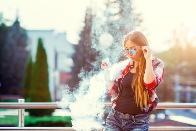 Photo young pretty hipster woman vaping and releases a cloud of vapor the evening sunset over the city ton