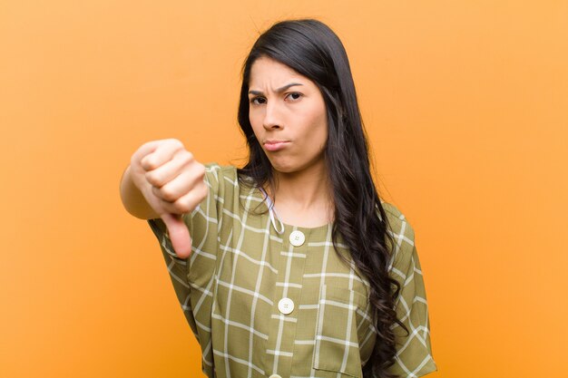 Young pretty hispanic woman feeling cross, angry, annoyed, disappointed or displeased, showing thumbs down with a serious look against brown wall