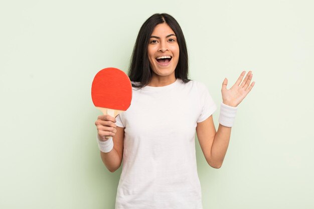 Young pretty hispanic woman feeling happy and astonished at something unbelievable ping pong concept