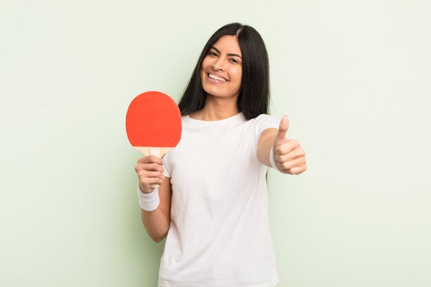 Young pretty hispanic woman feeling proudsmiling positively with thumbs up ping pong concept