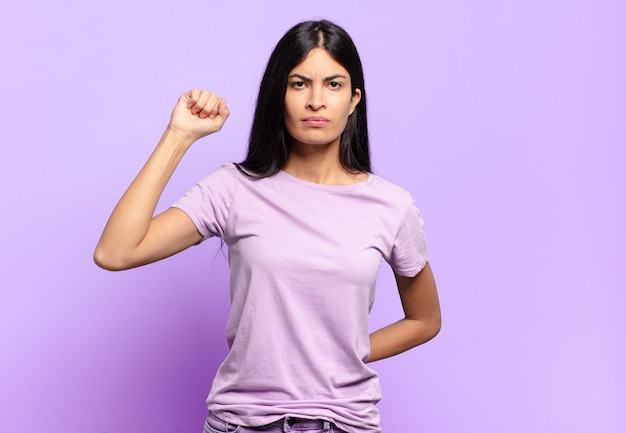 Young pretty hispanic woman feeling serious, strong and rebellious, raising fist up, protesting or fighting for revolution