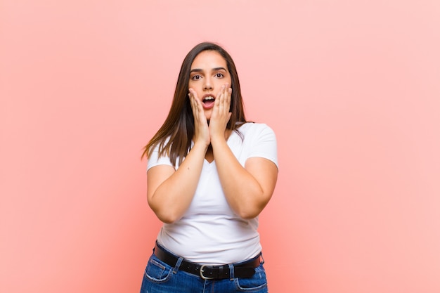 Young pretty hispanic woman feeling shocked and scared, looking terrified with open mouth and hands on cheeks against pink wall