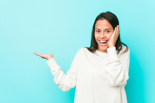 Young pretty hispanic woman holds copy space on a palm, keep hand over cheek. Amazed and delighted.