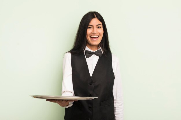 Young pretty hispanic woman looking happy and pleasantly surprised waiter with tray concept