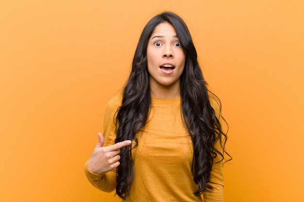 Young pretty hispanic woman looking shocked and surprised with mouth wide open, pointing to self against brown wall