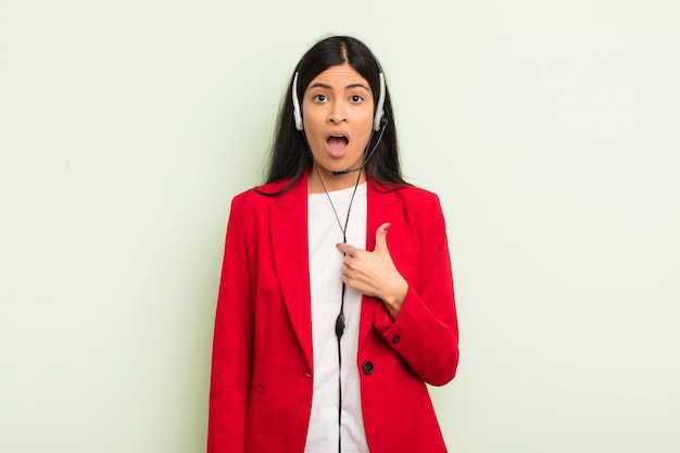 young pretty hispanic woman looking shocked and surprised with mouth wide open, pointing to self. telemarteker concpet