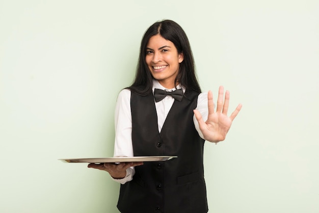 Young pretty hispanic woman smiling and looking friendly showing number five waiter with tray concept
