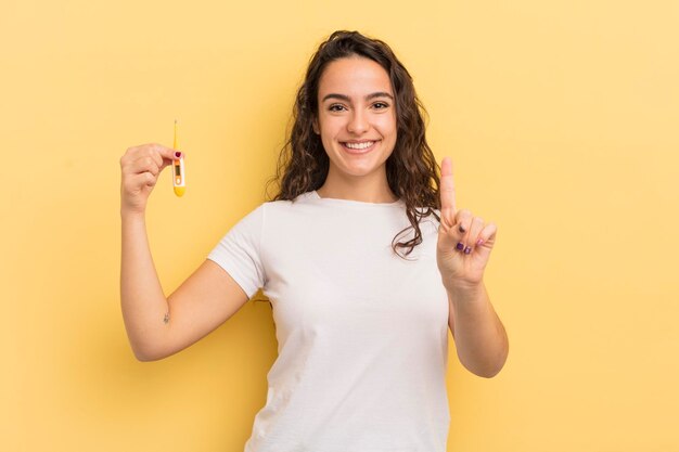 young pretty hispanic woman smiling and looking friendly, showing number one. thermometer concept