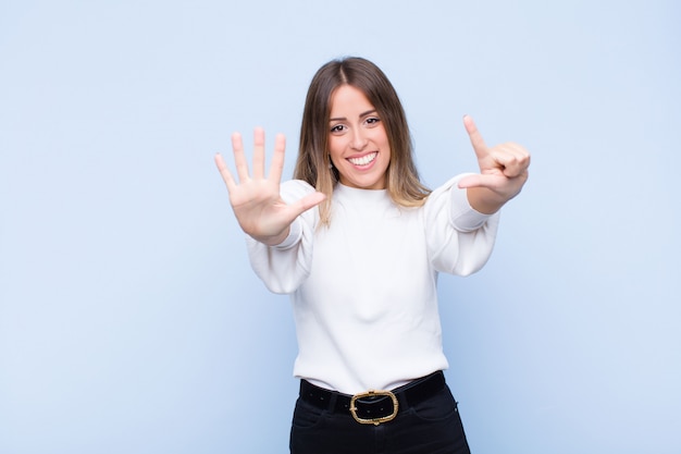 Young pretty hispanic woman smiling and looking friendly, showing number seven or seventh with hand forward, counting down against blue wall