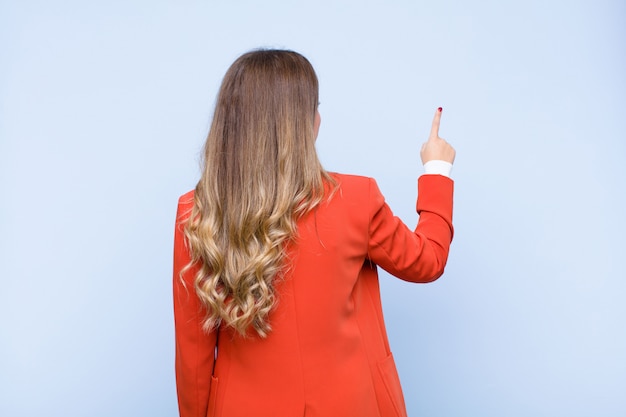 Young pretty hispanic woman standing and pointing to object on copy space, rear view over blue wall