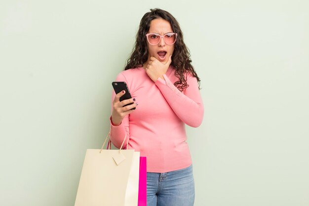 Young pretty hispanic woman with mouth and eyes wide open and hand on chin shopping bags concept