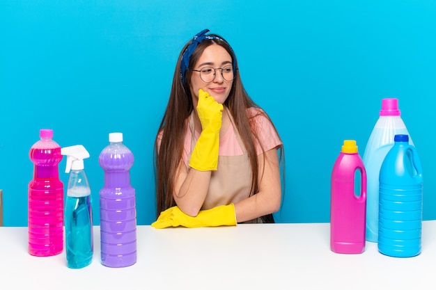 Young pretty house keeper smiling with a happy, confident expression with hand on chin, wondering and looking to the side