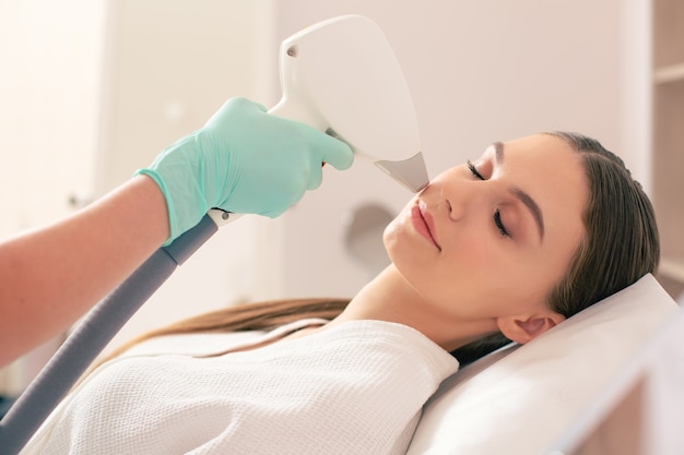 Young pretty lady having her eyes closed while experienced beautician holding a special laser and removing hair on the face of woman