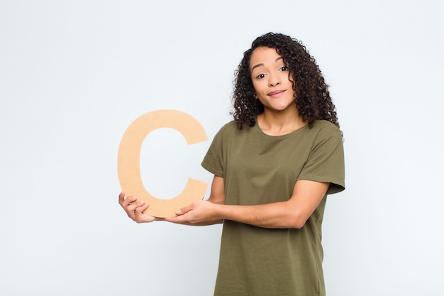 Young pretty latin woman confused, doubtful, thinking, holding the letter C of the alphabet to form a word or a sentence.