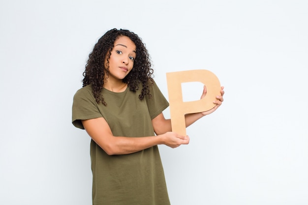 Young pretty latin woman confused, doubtful, thinking, holding the letter P of the alphabet to form a word or a sentence.