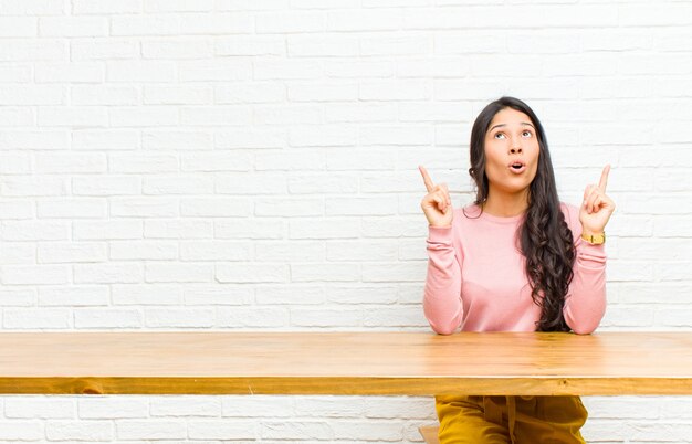 Молодая милая латинская женщина, чувствуя трепет и открытый рот, указывая наверх с потрясенным и удивленным взглядом, сидя перед столом