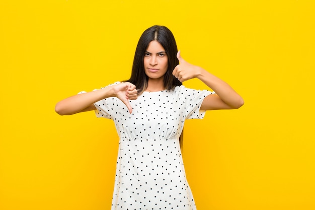 Young pretty latin woman feeling confused, clueless and unsure, weighting the good and bad in different options or choices over wall