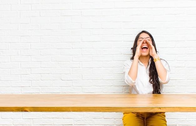 Young  pretty latin woman feeling happy, excited and positive, giving a big shout out with hands next to mouth, calling out sitting in front of a table