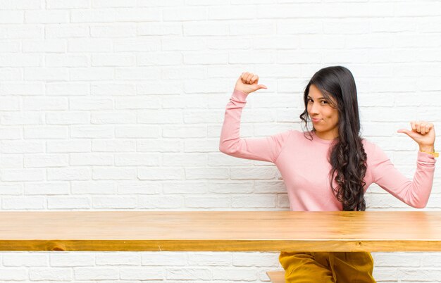 Young  pretty latin woman feeling proud, arrogant and confident, looking satisfied and successful, pointing to self sitting in front of a table