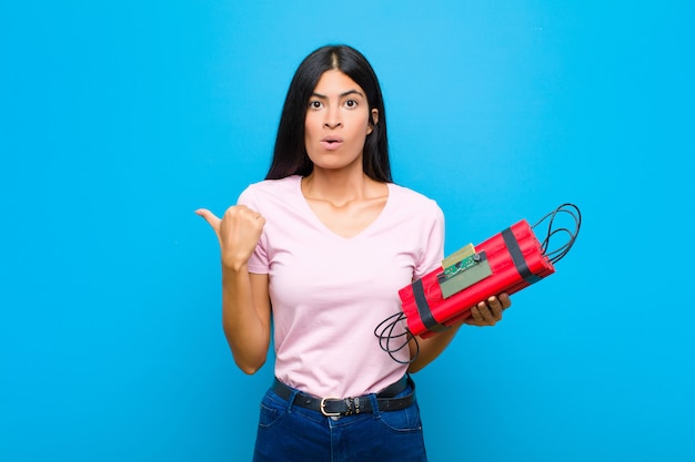 Young pretty latin woman looking astonished in disbelief, pointing at object on the side and saying wow, unbelievable against flat wall