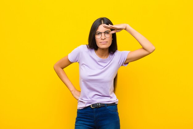 Young pretty latin woman looking bewildered and astonished, with hand over forehead looking far away, watching or searching against flat wall