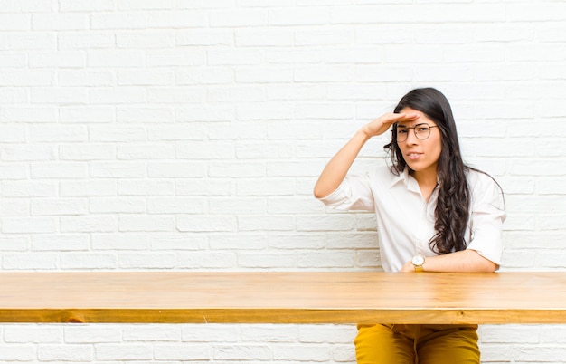 Young  pretty latin woman looking bewildered and astonished, with hand over forehead looking far away, watching or searching sitting in front of a table