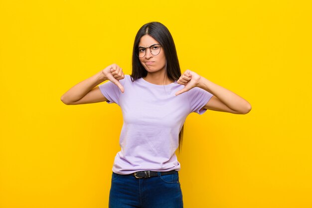 Young pretty latin woman looking sad, disappointed or angry, showing thumbs down in disagreement, feeling frustrated against flat wall