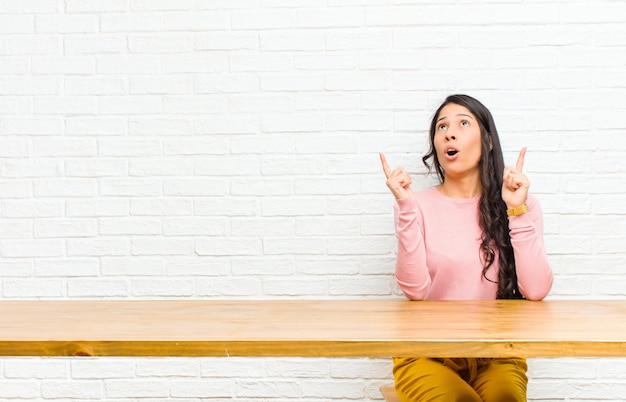 Young  pretty latin woman looking shocked, amazed and open mouthed, pointing upwards with both hands to copyspace sitting in front of a table