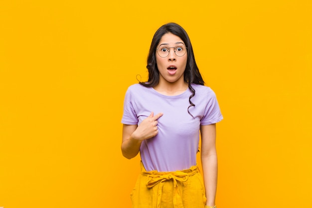 Young pretty latin woman looking shocked and surprised with mouth wide open, pointing to self against orange wall