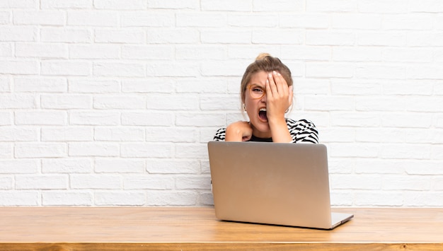 Young pretty latin woman looking sleepy, bored and yawning, with a headache and one hand covering half the face sitting with her laptop