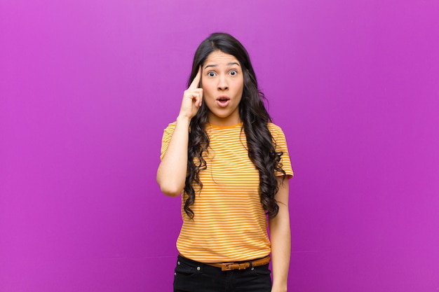 Young pretty latin woman looking surprised, open-mouthed, shocked, realizing a new thought, idea or concept against purple wall