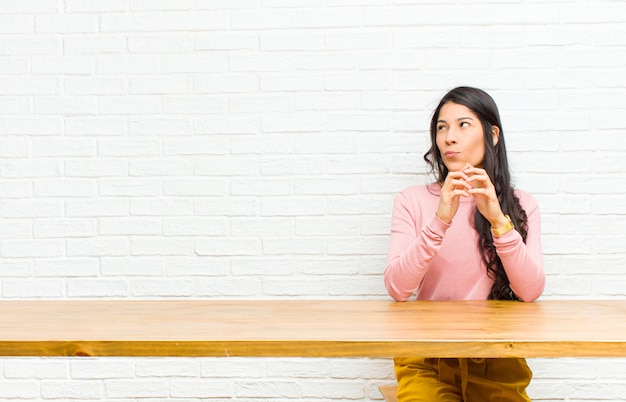 Photo young  pretty latin woman scheming and conspiring, thinking devious tricks and cheats, cunning and betraying sitting in front of a table