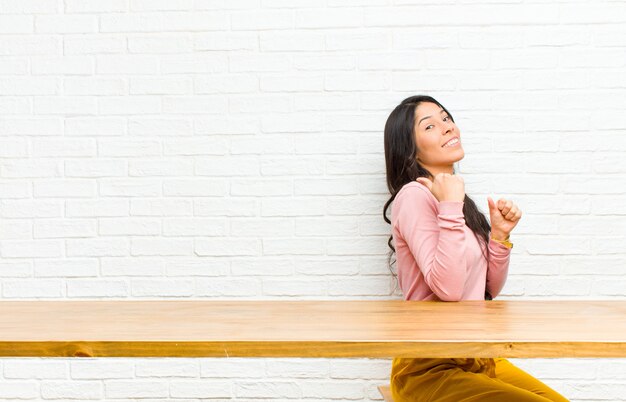 Young  pretty latin woman smiling cheerfully and casually pointing to copy space on the side, feeling happy and satisfied sitting in front of a table