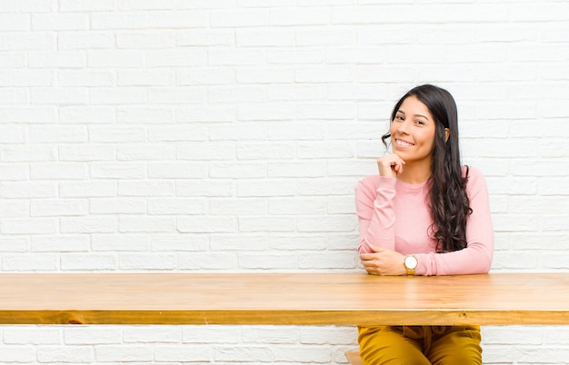 Young  pretty latin woman smiling, enjoying life, feeling happy, friendly, satisfied and carefree with hand on chin sitting in front of a table