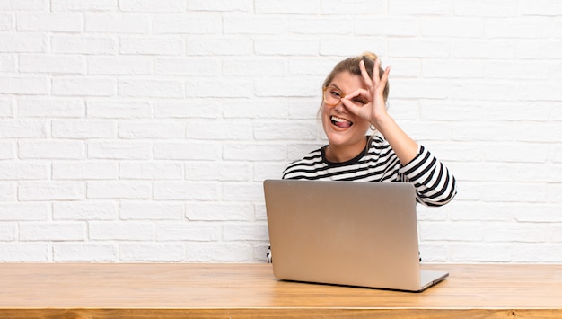 Young pretty latin woman smiling happily with funny face, joking and looking through peephole, spying on secrets sitting with her laptop