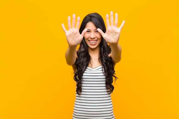 Young pretty latin woman smiling and looking friendly, showing number ten or tenth with hand forward, counting down against orange wall