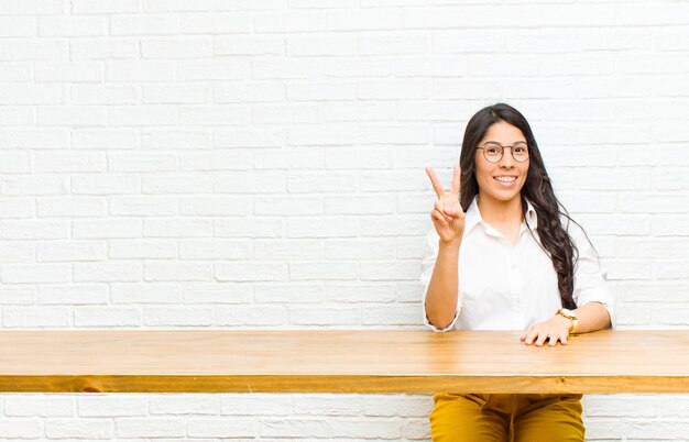 Young  pretty latin woman smiling and looking friendly, showing number two or second with hand forward, counting down sitting in front of a table