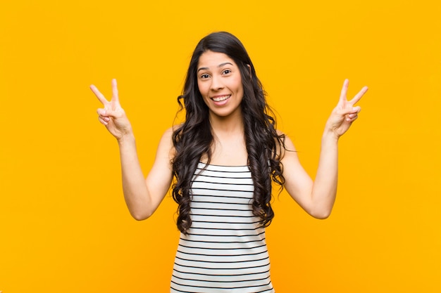 Young pretty latin woman smiling and looking happy, friendly and satisfied, gesturing victory or peace with both hands against orange wall