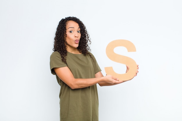 Young pretty latin woman surprised, shocked, amazed, holding the letter S of the alphabet to form a word or a sentence.