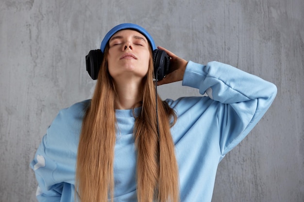 파란색 스웨터에 재미있는 모자와 검은색 헤드폰을 쓴 젊은 꽤 긴 머리 소녀가 음악을 듣습니다.